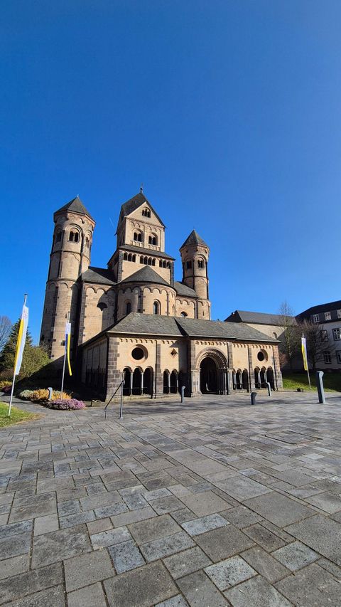Eine beeindruckende Kirche mit drei Türmen vor klarem, blauem Himmel. Der Platz vor der Kirche ist mit Steinen gepflastert und es gibt einige Flaggen.