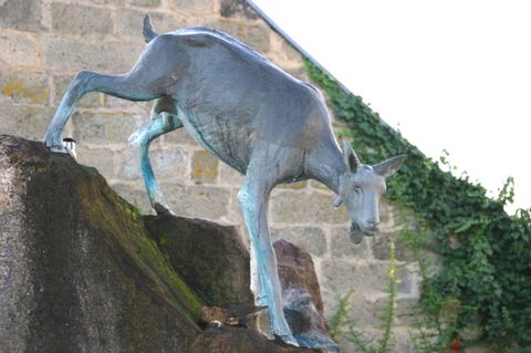 Eine Statue einer ziegenartigen Figur, die auf einem Felsen steht. Die Skulptur ist aus Bronze und hat einen metallic-blauen Farbton.