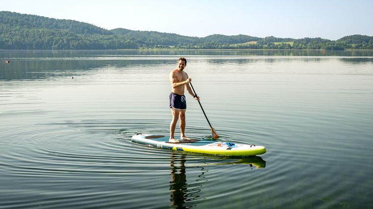 Eine Person steht auf einem Stand-Up-Paddle-Board in einem ruhigen See. Im Hintergrund sind sanfte Hügel und ein klarer Himmel zu sehen.