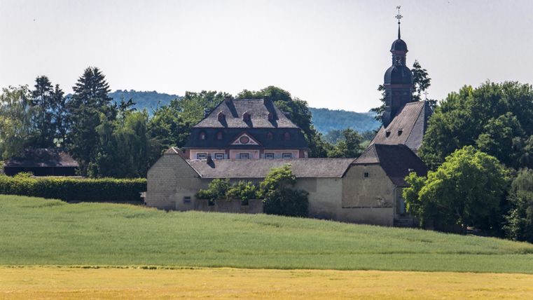 Ein charmantes Landhaus umgeben von Wiesen und Bäumen. Im Hintergrund ragt ein Kirchturm in den Himmel.