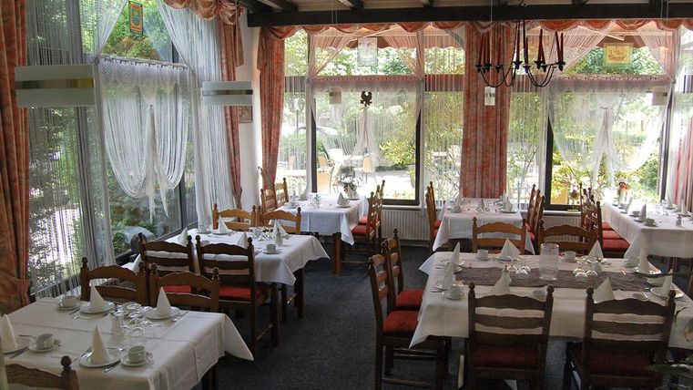 Ein gemütliches Restaurant mit gedeckten Tischen und Holzstühlen. Große Fenster sorgen für viel Tageslicht und einen Blick auf den Garten.