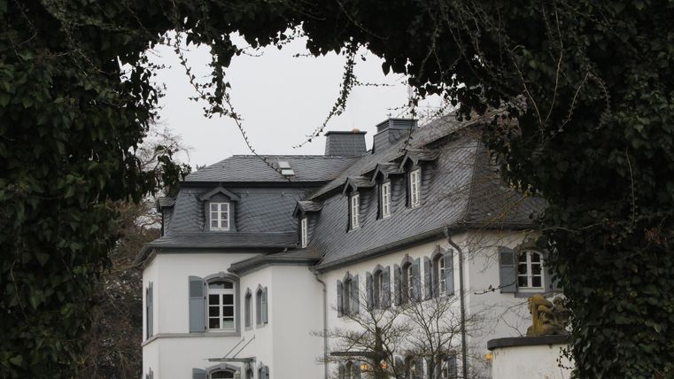 Ein elegantes, weißes Gebäude, umgeben von Bäumen. Im Vordergrund ist ein grüner Rahmen zu sehen.