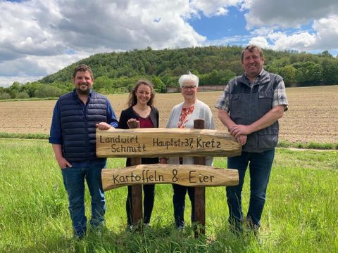 Eine Gruppe von vier Personen steht vor einem Holzschild mit den Aufschriften "Landwirt Schmitt" und "Kartoffeln & Eier". Im Hintergrund sieht man ein Feld und bewaldete Hügel unter einem wolkigen Himmel.