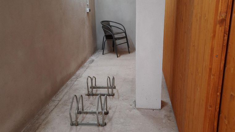 Ein schmaler Flur mit Wänden in neutrale Farbtönen. Am Boden stehen zwei Metallgestelle und ein Stuhl ist an der Wand platziert.