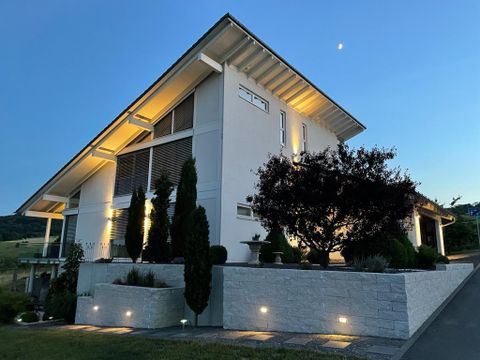 Une maison moderne avec une façade élégante et un éclairage extérieur. Le jardin est soigné et décoré d'arbres et de buissons.