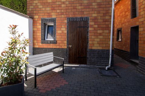 Eine kleine Terrasse mit einem grauen Bank und einer Pflanze. Im Hintergrund sieht man eine rustikale Wand aus Ziegelsteinen und eine Holztür.