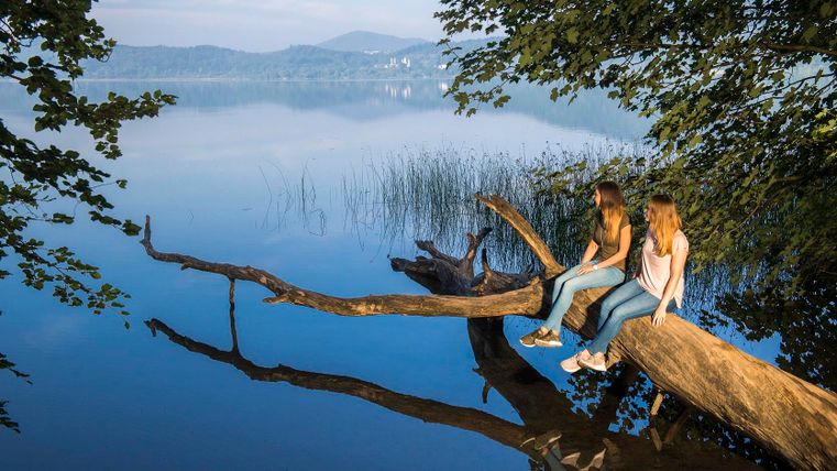 Zwei Mädchen sitzen auf einem Baumstamm über einem ruhigen See. Der Himmel ist klar und das Wasser spiegelt die Umgebung wider.