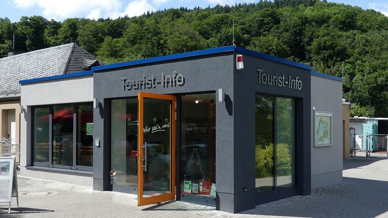 Ein modernes Tourist-Info-Gebäude mit großen Fenstern und einer einladenden Eingangstür. Es liegt in einer grünen Umgebung, umgeben von Bäumen.