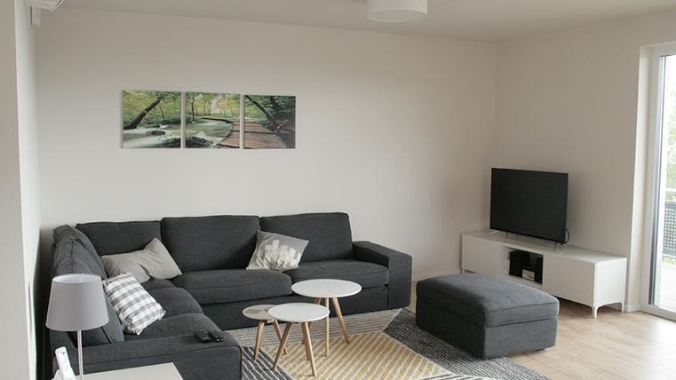Ein modernes Wohnzimmer mit einem grauen Sofa und einem Fernseher. Der Raum ist hell und gemütlich mit einem Teppich und kleinen Tischen.