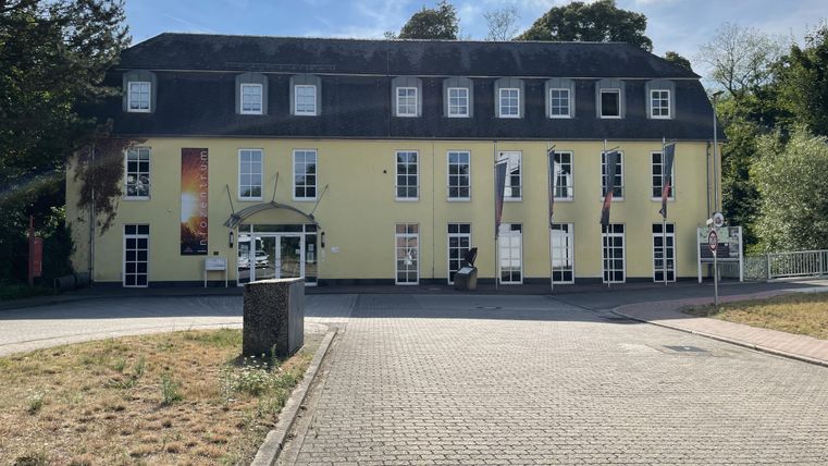Ein gelbes Gebäude mit mehreren Fenstern und einem Satteldach. Im Vordergrund ist ein gepflasterter Platz und etwas Grünbereich sichtbar.