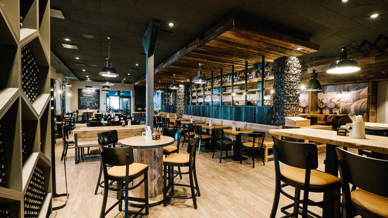 Ein modernes Restaurant mit Holzdekor und einer einladenden Atmosphäre. Die Einrichtung umfasst Tische und Stühle sowie eine lange Bar im Hintergrund.