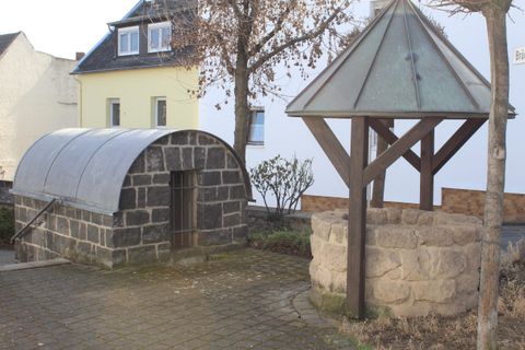 Ein historisches Steingebäude mit einem gewölbten Dach steht neben einem Brunnen mit einem Holzüberdach. Die Umgebung ist schlichten, mit Bäumen und Wohnhäusern.