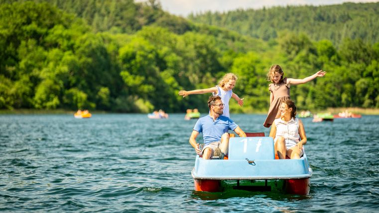 A family is riding on a pedal boat on a tranquil lake. In the background, green trees and other boaters are visible.