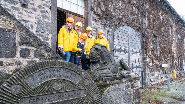 A group of five people stands in yellow rain jackets and helmets in front of an old building covered in ivy. Next to them is a stone with the image of a lion.