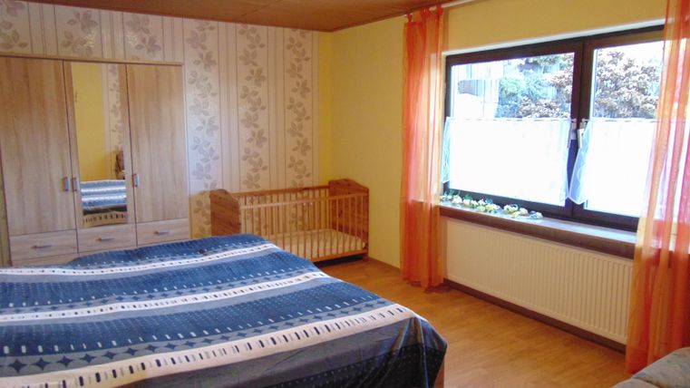 A simple bedroom with a large bed and a cozy armchair. The room has wooden flooring and a windowsill with plants.