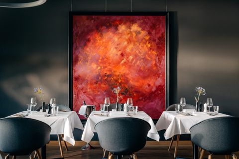 Ein elegantes Restaurant mit runden Tischen und hochwertigen Stühlen. An der Wand hängt ein großes, abstraktes Gemälde in warmen Rottönen.