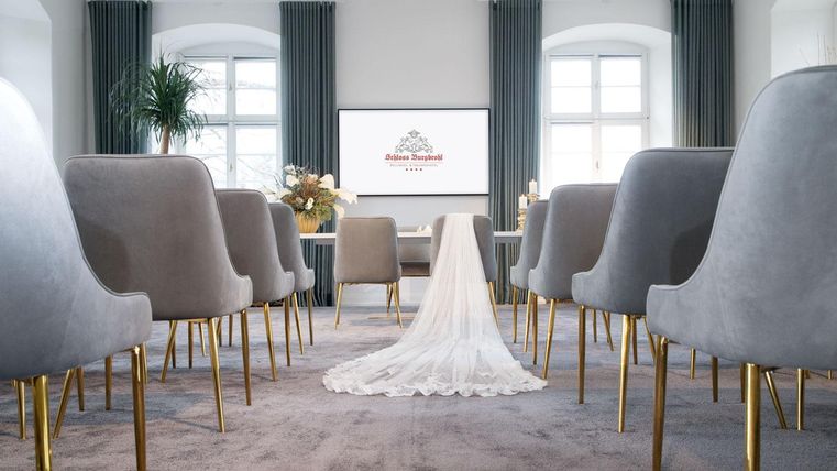 An elegant event space with gray chairs and a carpet. In the center lies a wedding dress, while a screen is visible in the background.