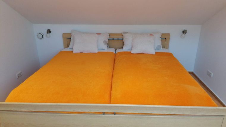A cozy bedroom with a double bed and orange bedcovers. The walls are plain, and there are reading lamps on both sides of the bed.