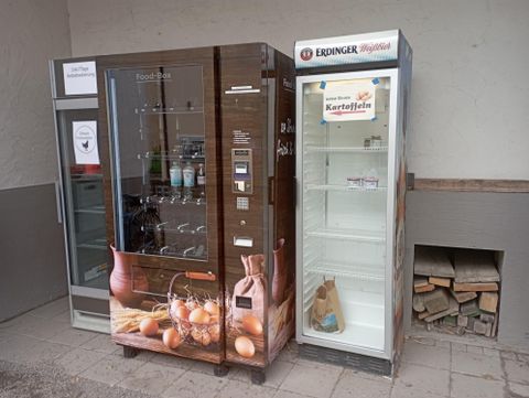 Ein Verkaufsautomat für Eier und Getränke steht neben einem leeren Kühlschrank für Kartoffeln. Beide sind an einer Wand aufgestellt.