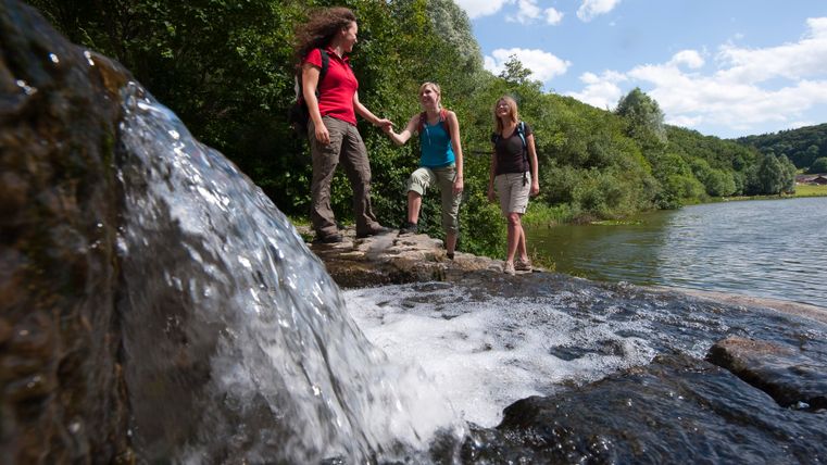 Drei Frauen wandern an einem Wasserfall in einem Waldgebiet entlang.