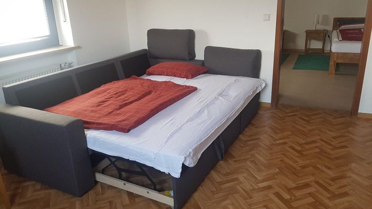 A sofa bed with red cushions and a blanket in a bright room. In the background, another room with a bed is visible.