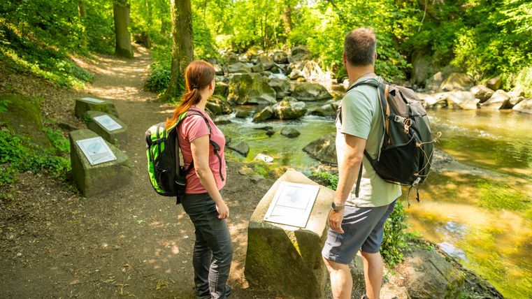 Zwei Wanderer stehen an einem kleinen Fluss in einem grünen Wald. Neben ihnen befinden sich Informationstafeln auf dem Weg.