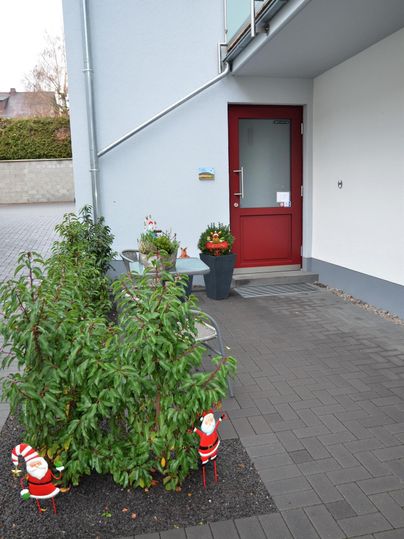 Ein Haus mit einer roten Tür und einer kleinen Eingangstreppe. Im Vordergrund stehen weihnachtliche Dekorationen und grüne Pflanzen.