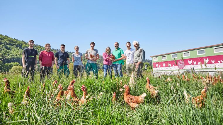 Eine Gruppe von Menschen steht auf einem grünen Feld, umgeben von Hühnern. Im Hintergrund sind ein Gebäude und ein klarer blauer Himmel zu sehen.