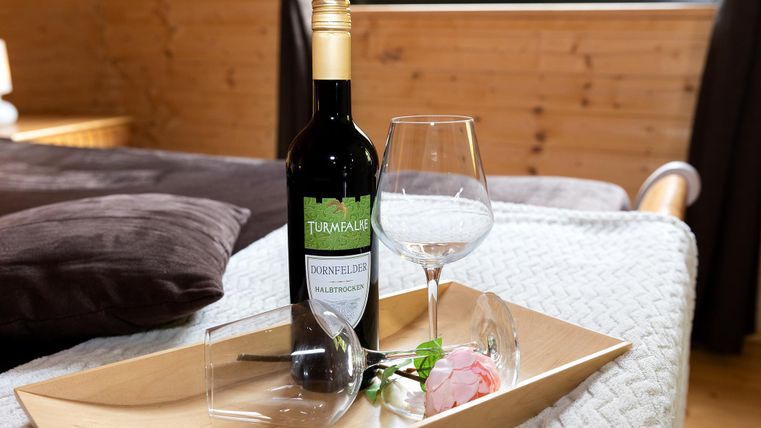 A bottle of red wine is next to a glass on a wooden tray. A pink rose and a transparent glass are also there.