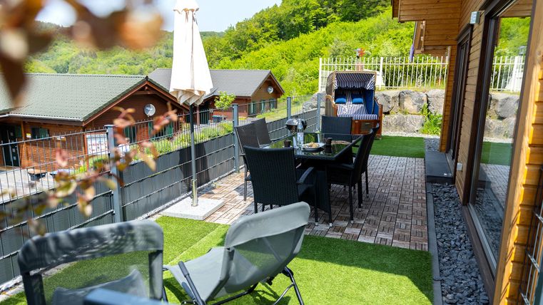 A cozy terrace with chairs and a table, surrounded by green landscape. In the background, holiday cabins are visible.