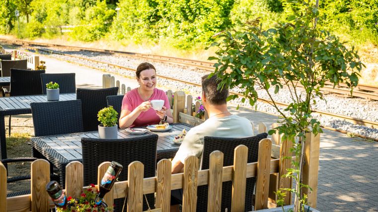 Ein gemütlicher Biergarten mit zwei Personen, die Kaffee und Essen genießen. Im Hintergrund sind grüne Bäume und Gleise zu sehen.