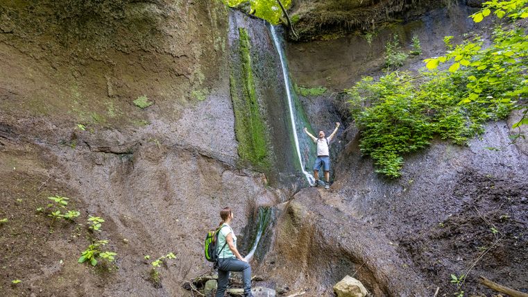 Zwei Personen stehen an einem kleinen Wasserfall in einer Schlucht, umgeben von Felsen und Pflanzen.