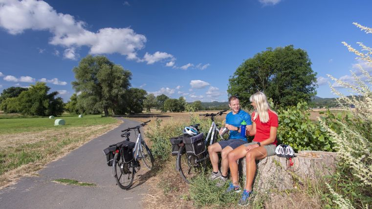 Ein Paar sitzt auf einem Baumstumpf neben einem Radweg. Im Hintergrund sind Bäume und ein klarer blauer Himmel zu sehen.