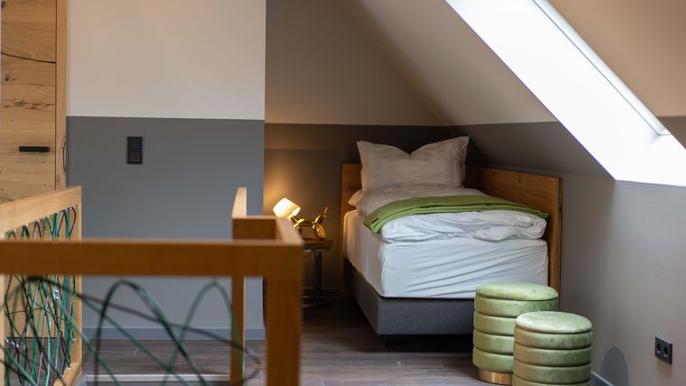 Ein modernes Zimmer mit einem Einzelbett und einer weißen Bettdecke. Zwei grüne Hocker stehen neben dem Bett, und das Licht schafft eine gemütliche Atmosphäre.