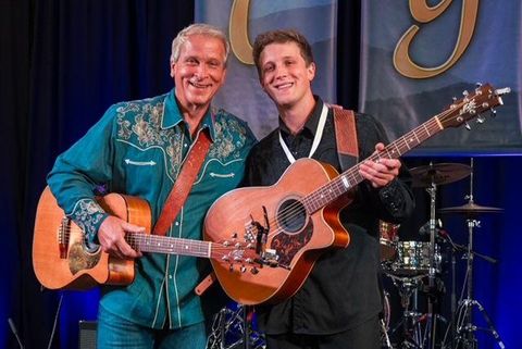 Zwei Musiker stehen auf einer Bühne und lächeln in die Kamera. Beide halten Akustikgitarren, während im Hintergrund eine Band zu sehen ist.