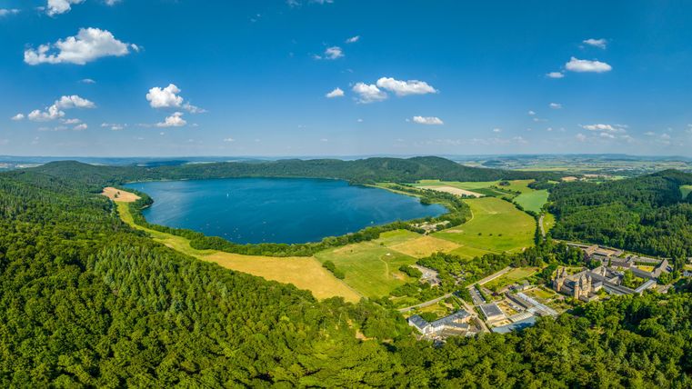 Ein schöner, klarer See umgeben von grünen Feldern und Bäumen. Im Hintergrund ist eine kleine Siedlung zu sehen.