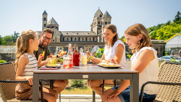 Eine Familie sitzt an einem Tisch im Freien und genießt ihr Essen. Im Hintergrund ist ein historisches Gebäude zu sehen.