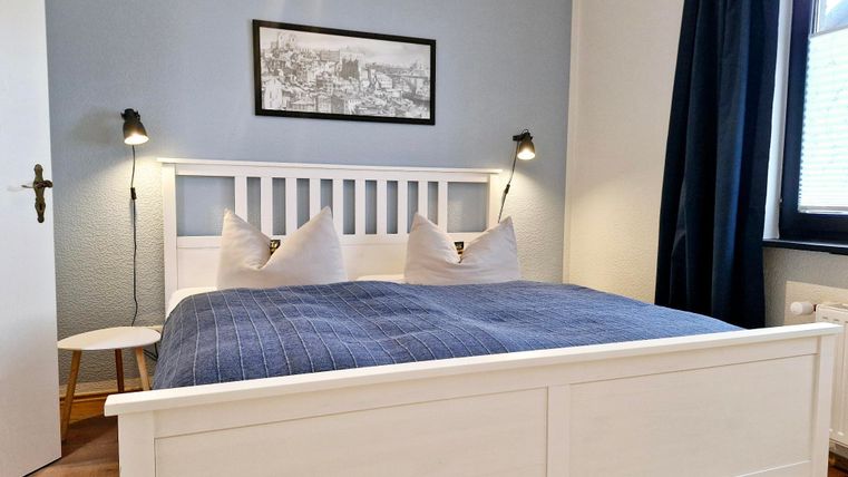 Ein gemütliches Schlafzimmer mit einem weißen Bett und einem blauen Bettüberwurf. Die Wände sind hellblau, und das Fenster hat einen Vorhang in dunklem Blau.
