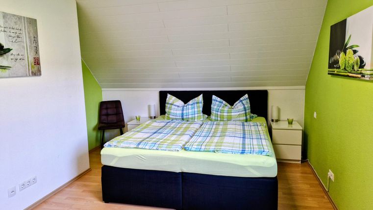 Ein modernes Schlafzimmer mit einem großen Bett und bunten Kissen. Die Wände sind in einem frischen Grünton gestrichen und es gibt einen Stuhl sowie dekorative Bilder.
