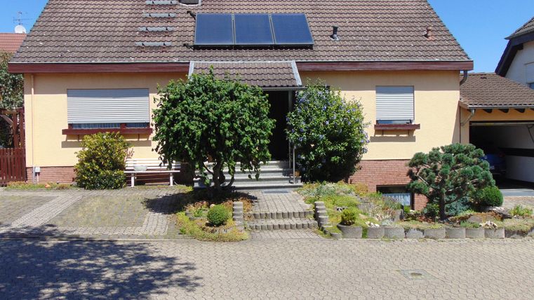 A family house with a reddish-brown roof and solar panels. The front yard is well-kept with various plants and shrubs.
