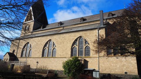 An impressive church with tall windows and a distinctive roof. The facade is made of light stone and is surrounded by trees.