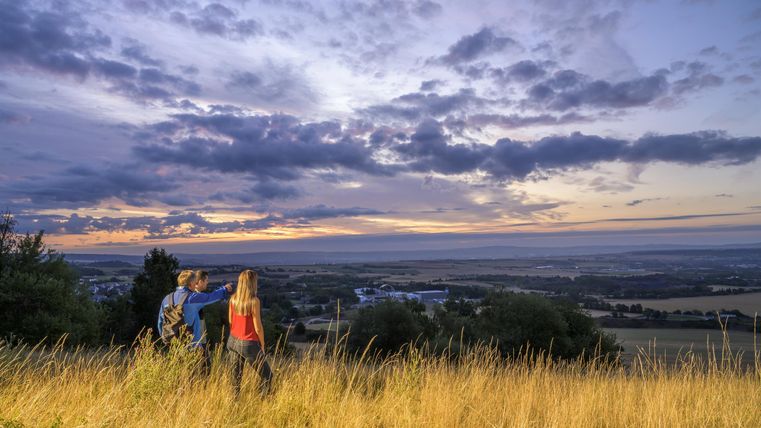 Zwei Personen stehen auf einer Wiese und schauen auf einen malerischen Sonnenuntergang. Der Himmel ist mit bunten Wolken bedeckt, während die Landschaft im Hintergrund zu sehen ist.