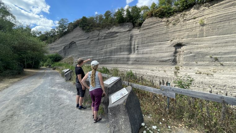 Zwei Personen stehen an einem Wanderweg und betrachten eine große Steinschicht. Der Himmel ist blau mit einigen Wolken, und die Umgebung ist grün und natürlich.