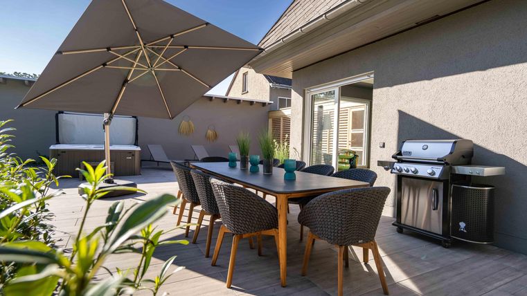 Ein gemütlicher Essbereich im Freien mit einem großen Tisch und Stühlen. Neben dem Tisch steht ein Grill und ein Sonnenschirm spendet Schatten.
