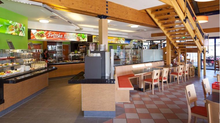 Ein modernes Restaurant mit einem freundlichen Ambiente. Ein Buffetbereich und Tische zum Essen sind sichtbar, sowie eine Holztreppe im Hintergrund.