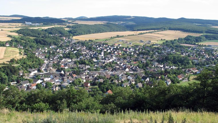 Eine ruhige Aussicht auf ein kleines Dorf, umgeben von grünen Hügeln und Feldern. Der Himmel ist klar und blau.