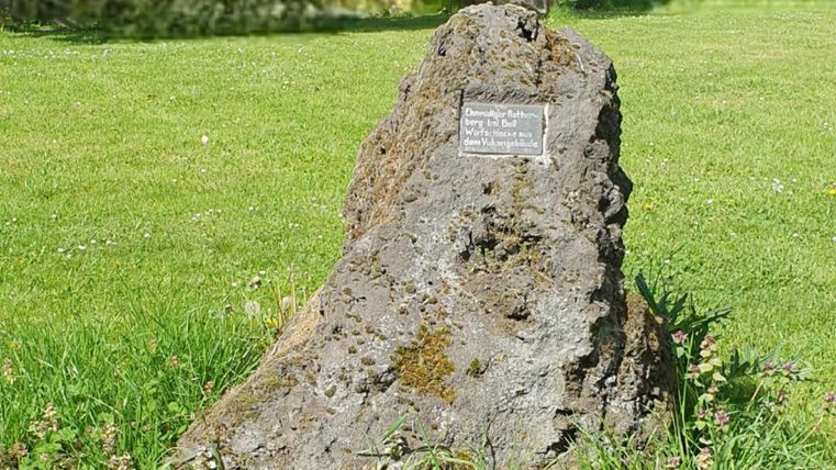 A large rock stands on a green meadow. A small plaque is attached to the rock.