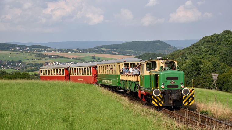 Ein nostalgischer Zug fährt durch eine grüne Landschaft. Die Wolken am Himmel sorgen für eine entspannte Atmosphäre.