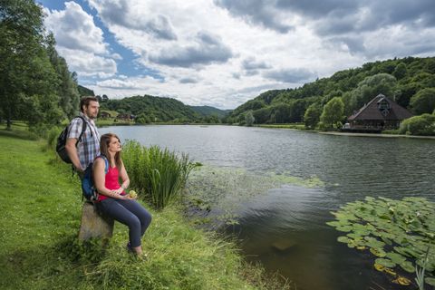 Zwei Personen sitzen am Ufer eines Sees mit bewaldeten Hügeln im Hintergrund.
