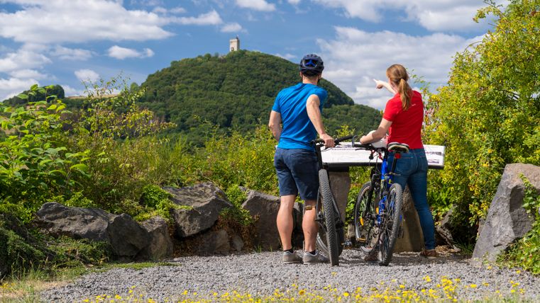 Zwei Fahrradfahrer stehen mit ihren Fahrrädern auf einem Schotterweg. Vor Ihnen eine weiße Infotafel. Im Hintergrund sieht man einen grün bewaldeten Hügel mit einer Burgruine.